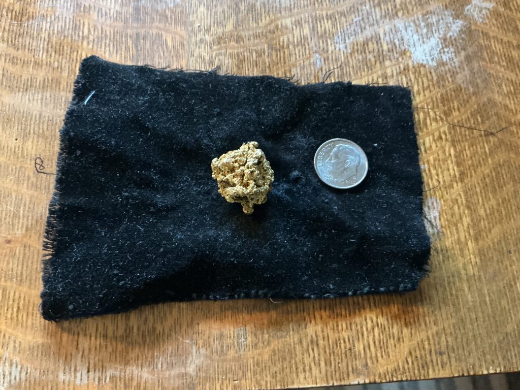 A single gold nugget on cloth next to a dime for scale